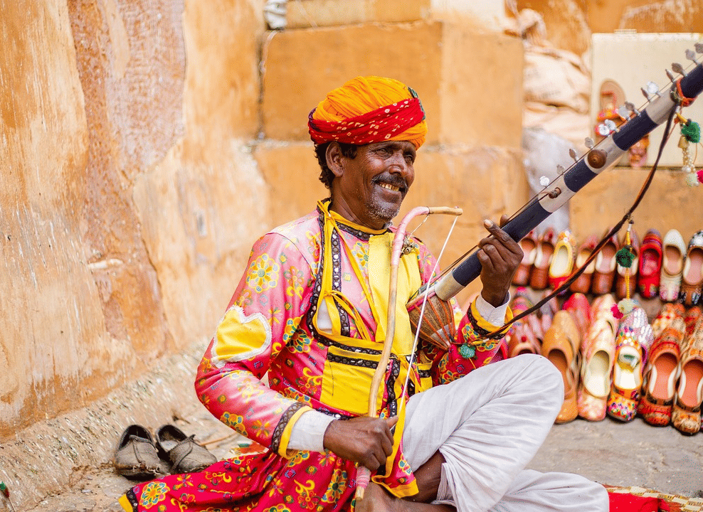 A musician in colourful traditional attire sits on the ground playing a stringed folk instrument, with rows of vibrant handcrafted shoes displayed behind him against an earthy wall.