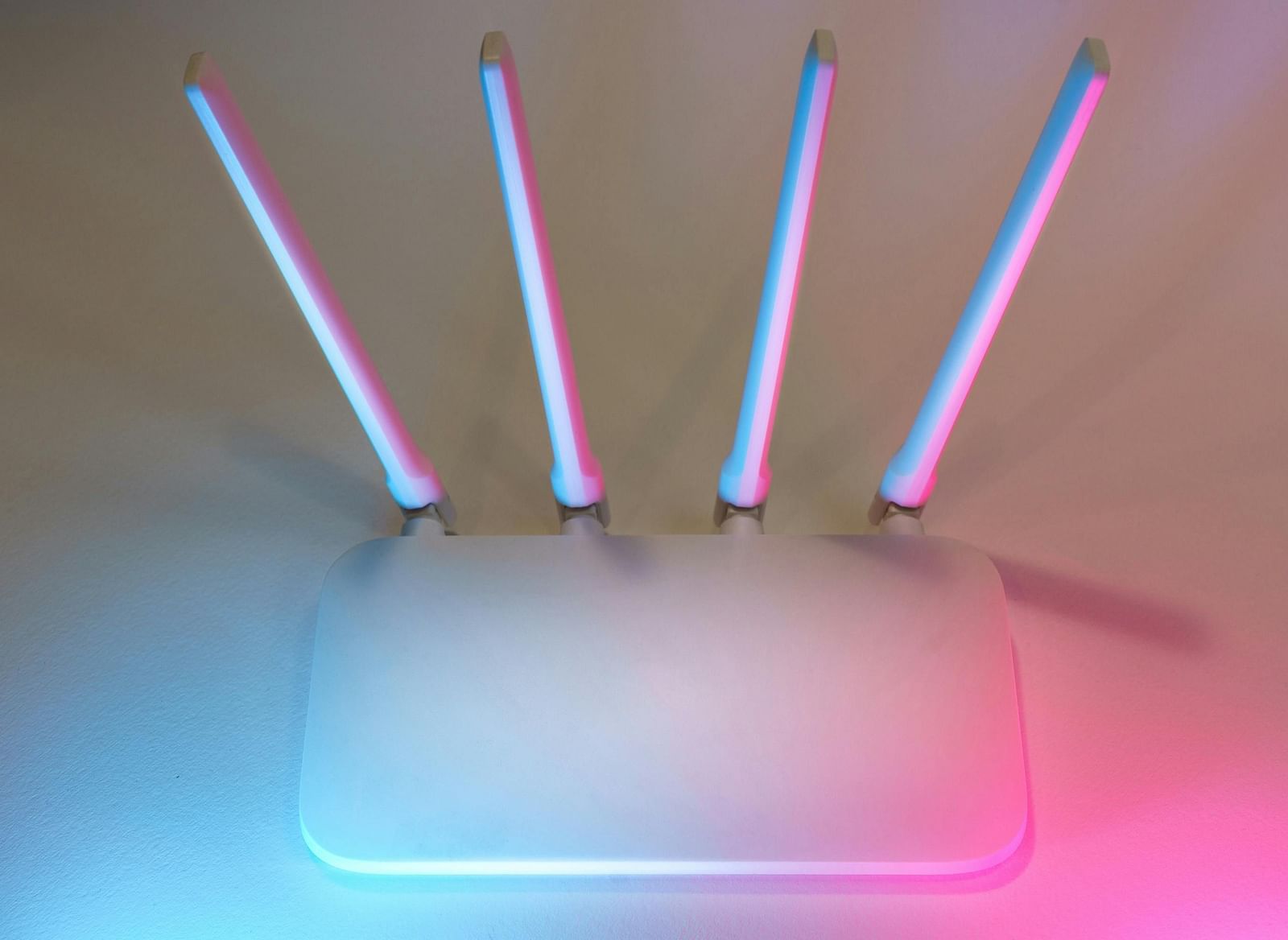 A white wireless router with four antennae illuminated in blue and pink hues against a neutral wall.