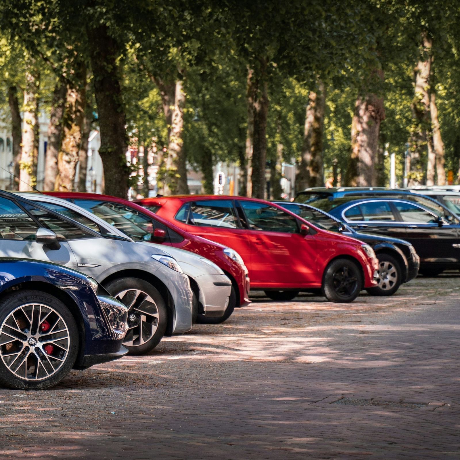 A row of parked cars in a shaded parking lot, with trees lining the sides and vehicles in various colours, in a well-maintained outdoor area.