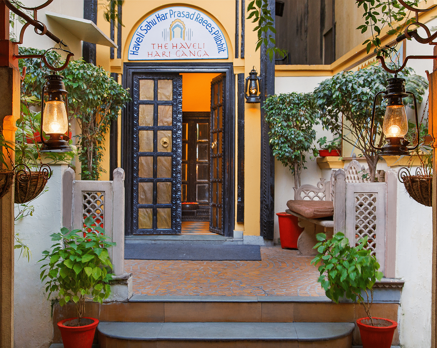 A wooden-furnished door at The Haveli Hari Ganga in Haridwar, adorned with plants, creates a warm and inviting entrance that enhances the beauty of the setting, welcoming guests with a touch of nature and charm.