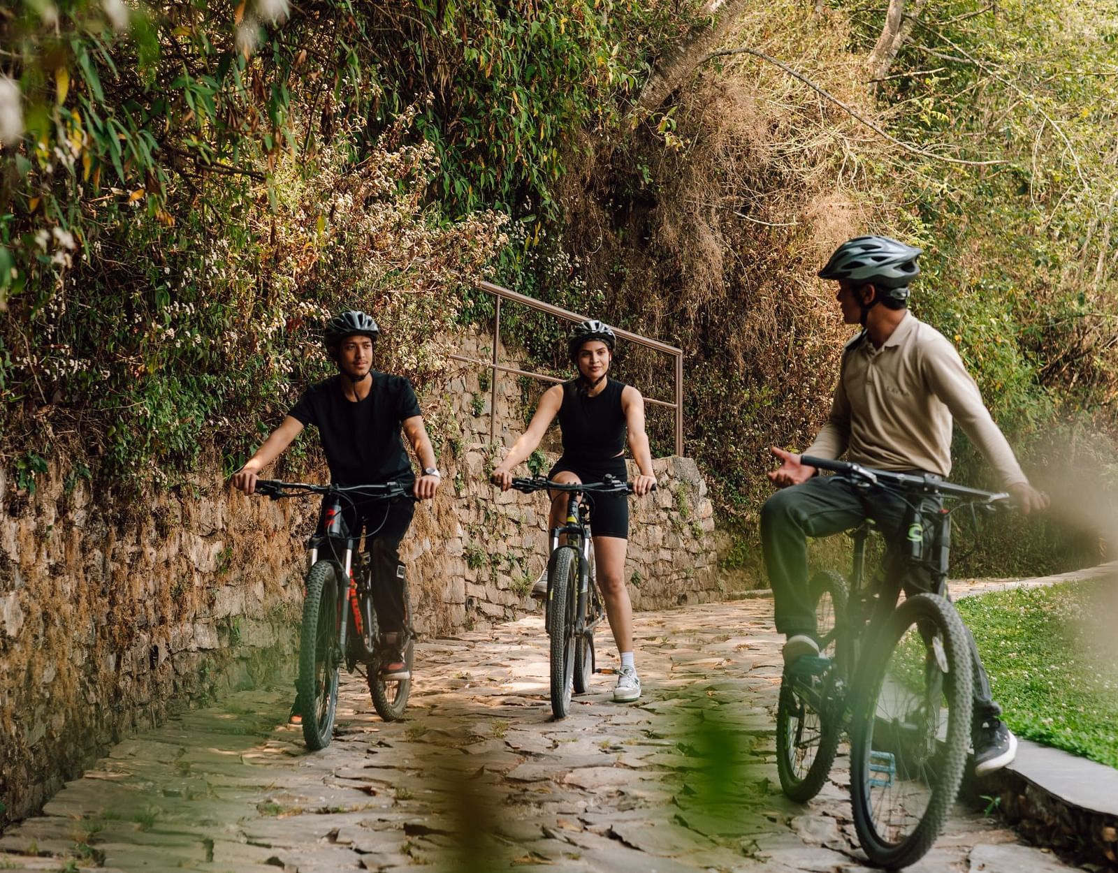 Three people cycling on a stone path surrounded by lush greenery, engaging in conversation.