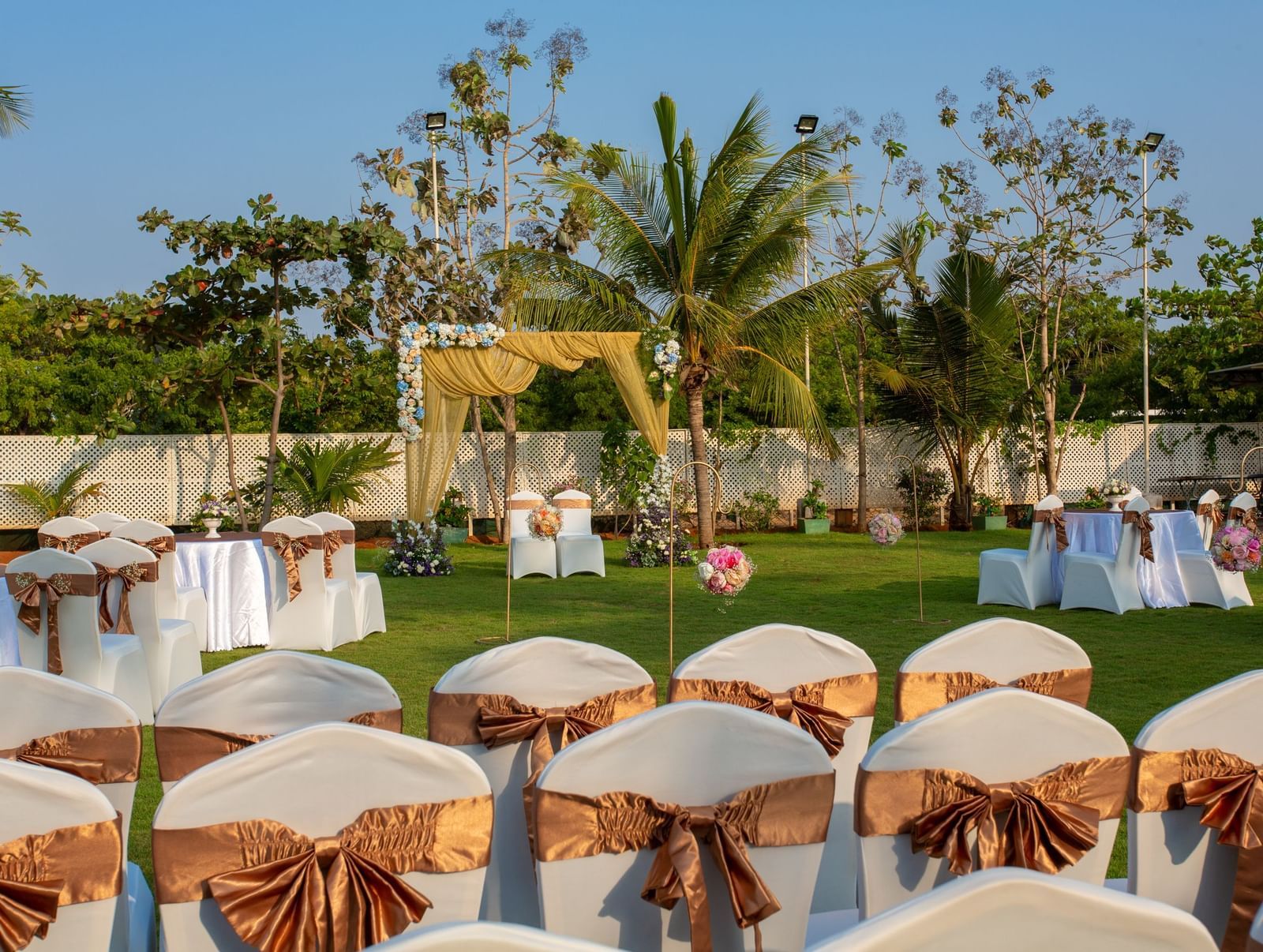 An outdoor event space with chairs covered in gold sashes, arranged on a green lawn, with a decorative archway in the distance | Grande Pavillion | Grand Bay Resort and Spa
