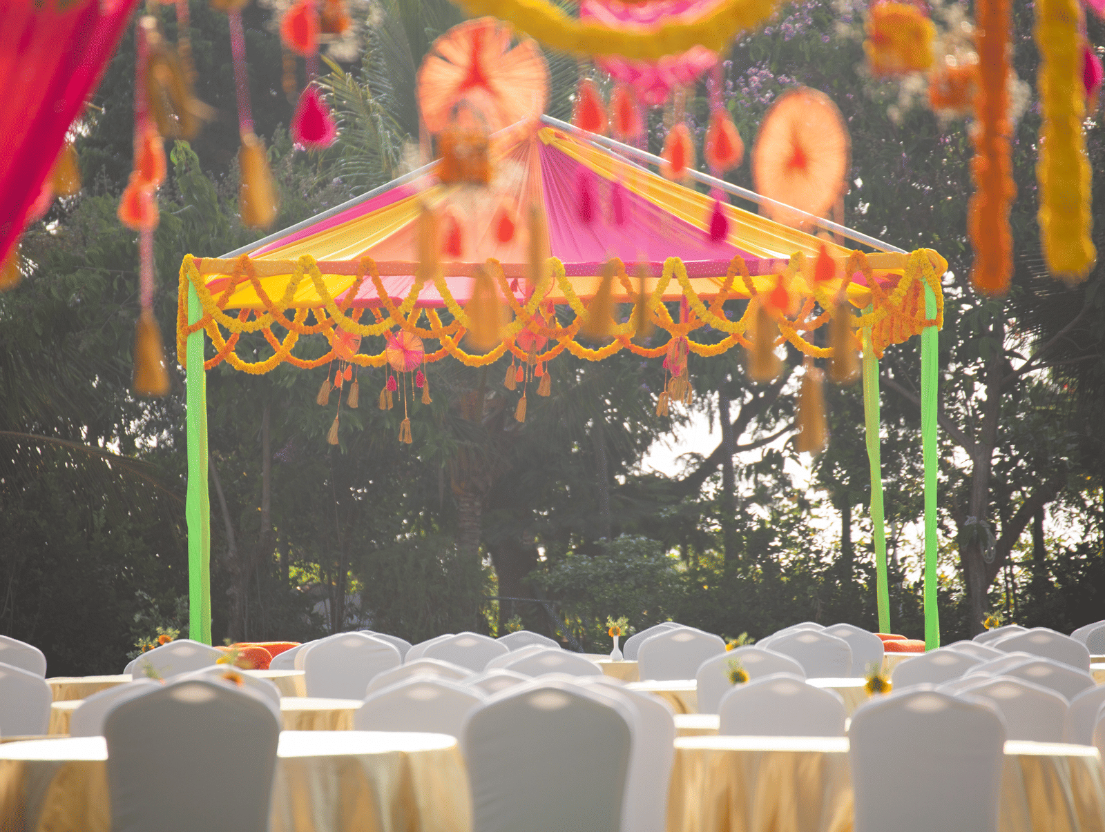 An outdoor wedding or event canopy in vibrant pink and yellow fabric, decorated with marigolds, set on a grassy lawn with round tables - Grande Bay Resort & Spa, Mamallapuram