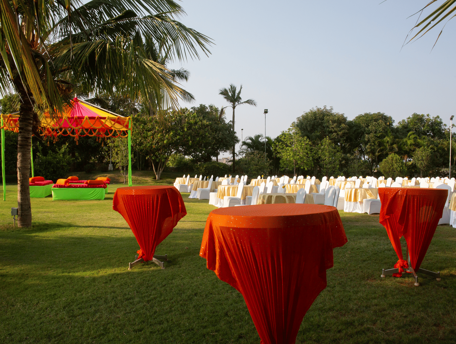 An outdoor gathering on a green lawn with tall orange cocktail tables and chairs, under the shade of palm trees - Grande Bay Resort & Spa, Mamallapuram