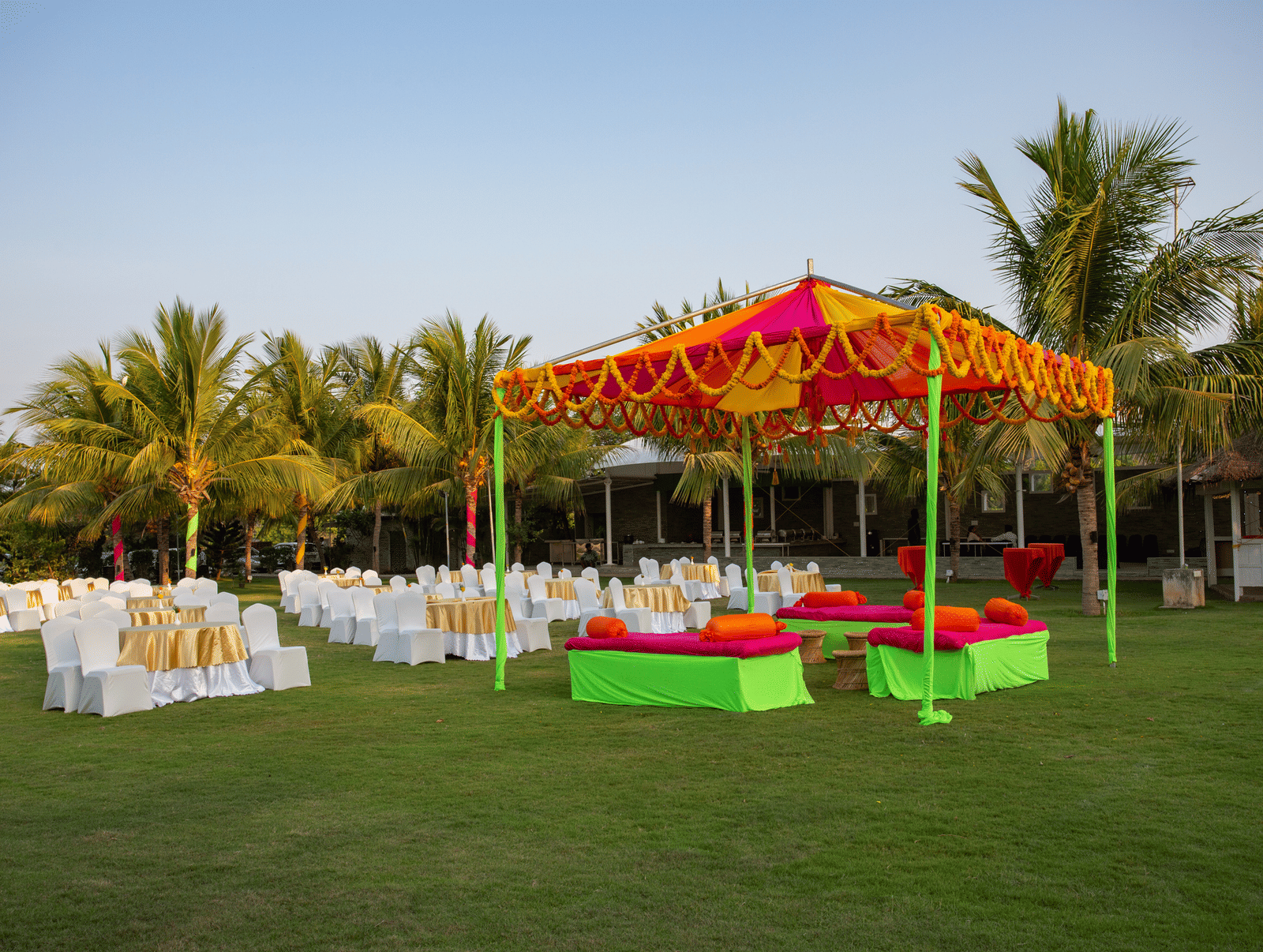 An outdoor event setup with an orange and yellow tent hosting green and red seating, and numerous white chairs on a grassy lawn - Grande Bay Resort & Spa, Mamallapuram