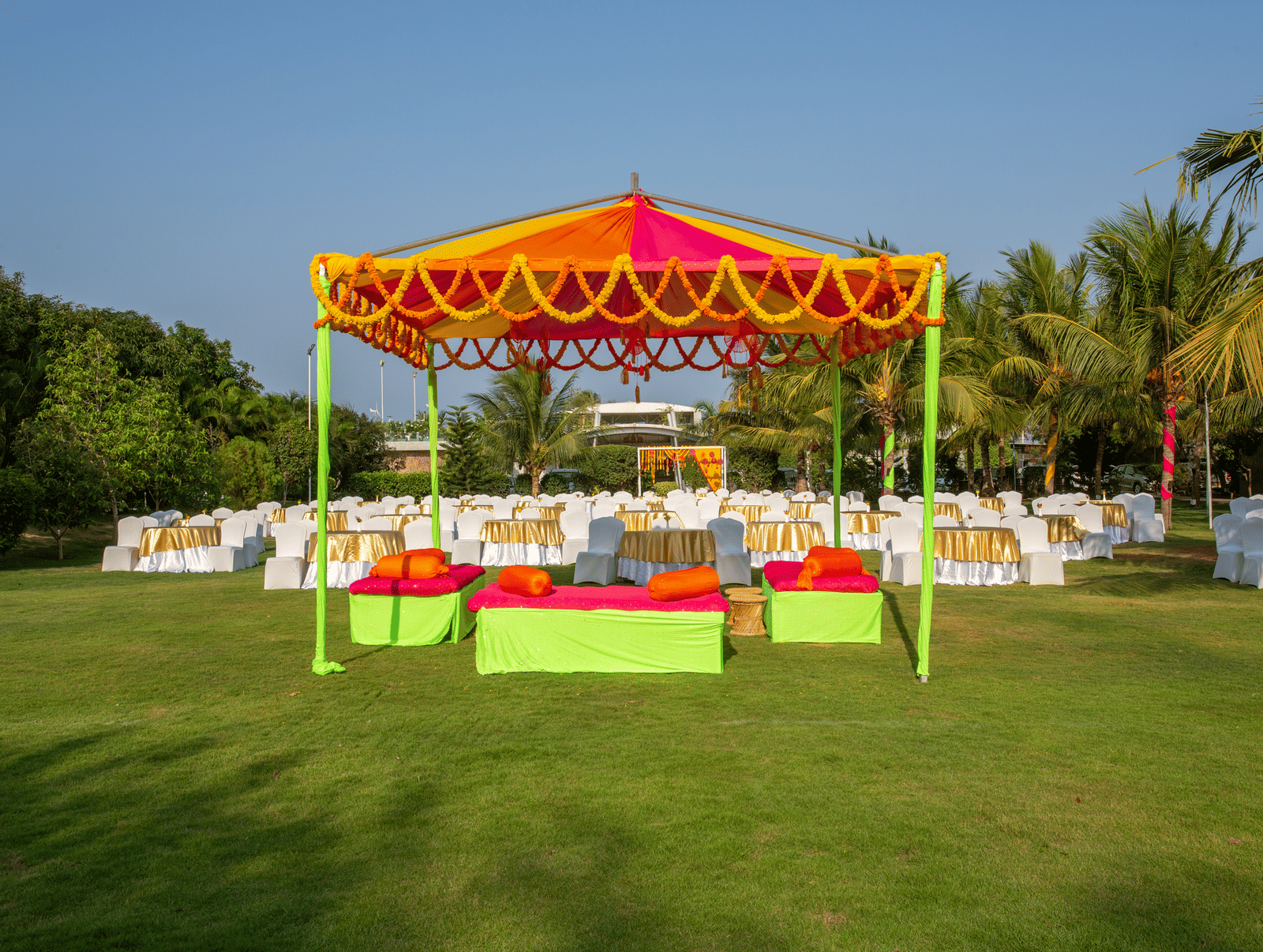 A colorful outdoor event setup featuring an orange and yellow tent, vibrant green and pink seating, and round tables on a manicured lawn - Grande Bay Resort & Spa, Mamallapuram