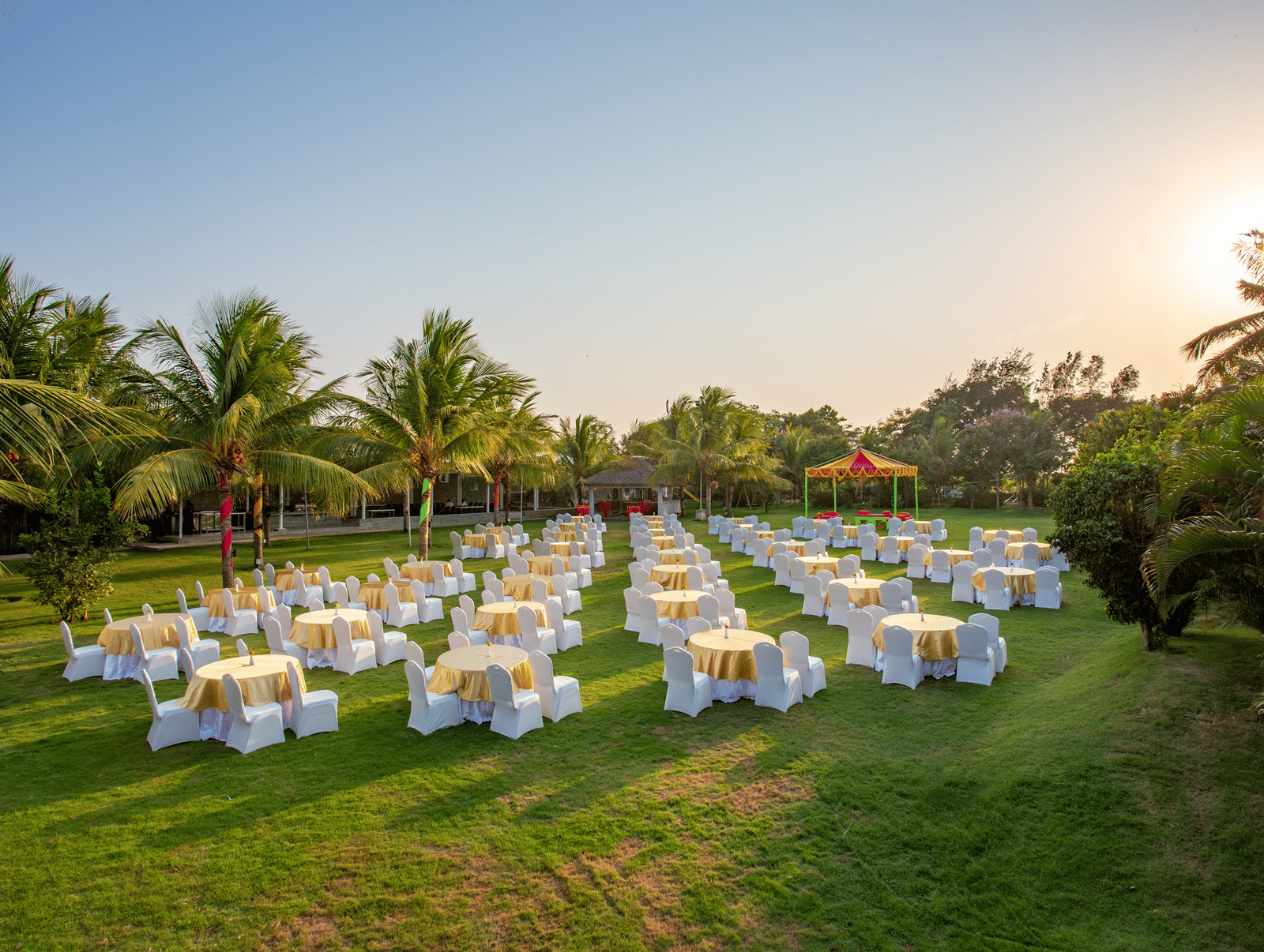 An outdoor event space on a green lawn with numerous round tables and chairs, set up for a gathering under a bright sky surrounded by palm trees - Grande Bay Resort & Spa, Mamallapuram