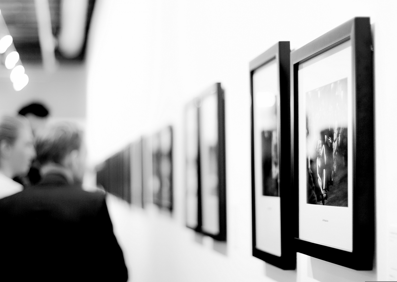 A black and white image of inside a museum with many paintings hung on the wall and people looking at them.
