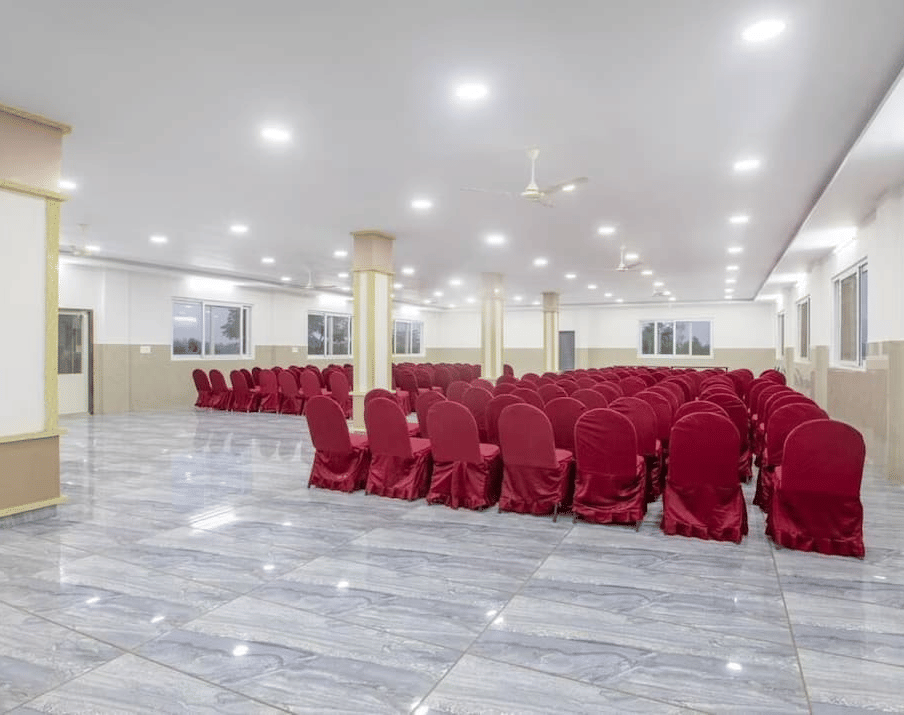 Back view of a spacious banquet hall at Siri Ambari Resorts and Spa, featuring red-covered chairs and bright lighting.