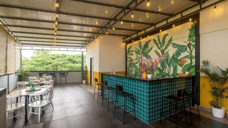 An outdoor, covered cafe area with a teal tiled bar, a tropical leaf mural, and white tables and chairs under warm string lights.