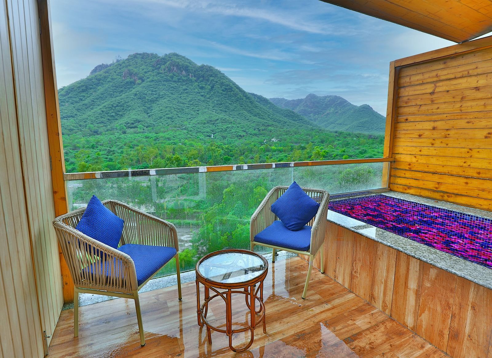 A balcony with two chairs, cushions on it, and a table with mountains as a scenery.- Bamboo Saa Resort & Spa, Udaipur