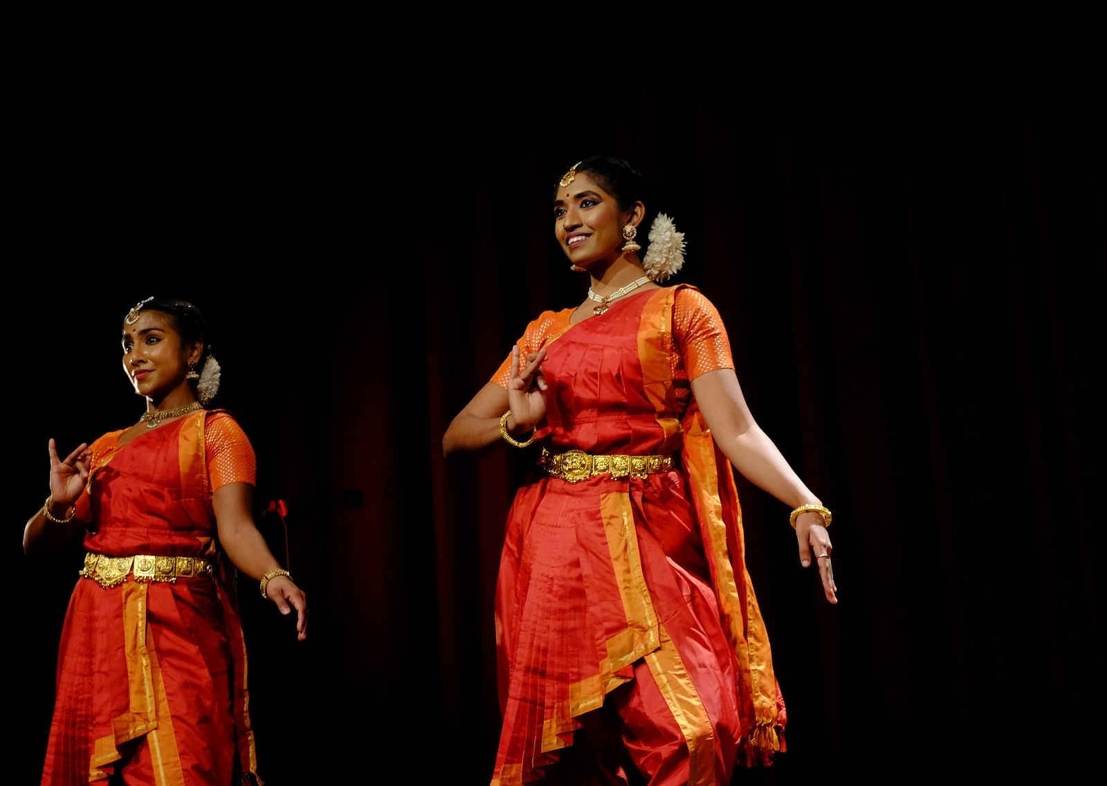 Two women performing bharatanatyam on stage with a black background.