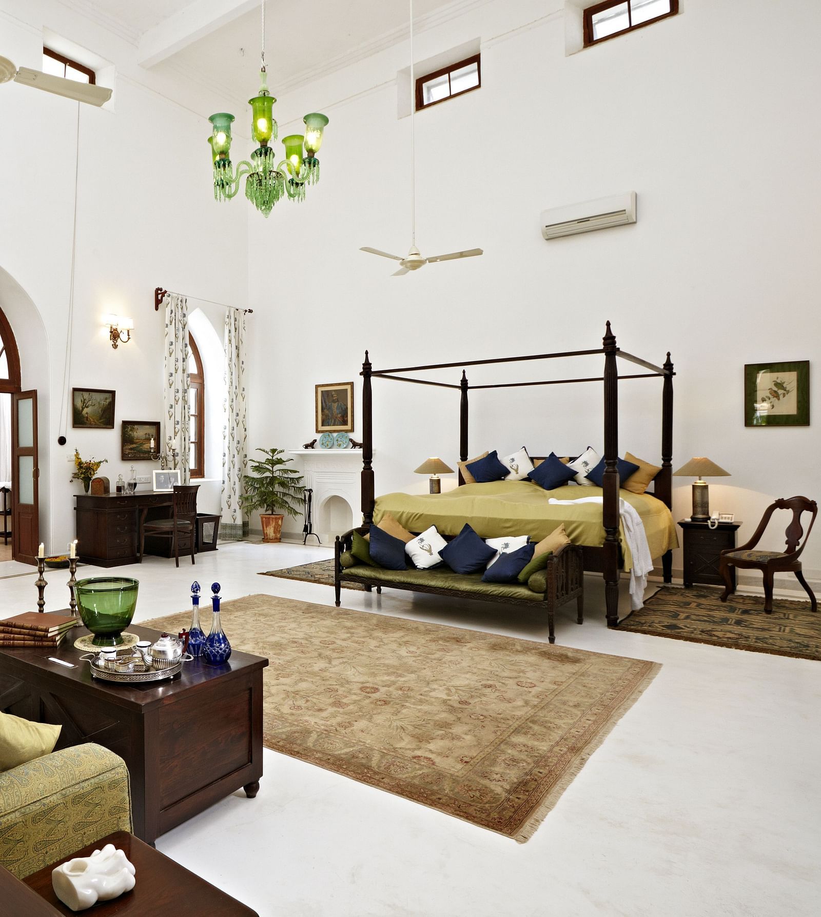 A spacious room with a four-poster bed, a sofa, and various furniture, featuring arched doorways and a high ceiling with fans and a chandelier- Shahpura Bagh.