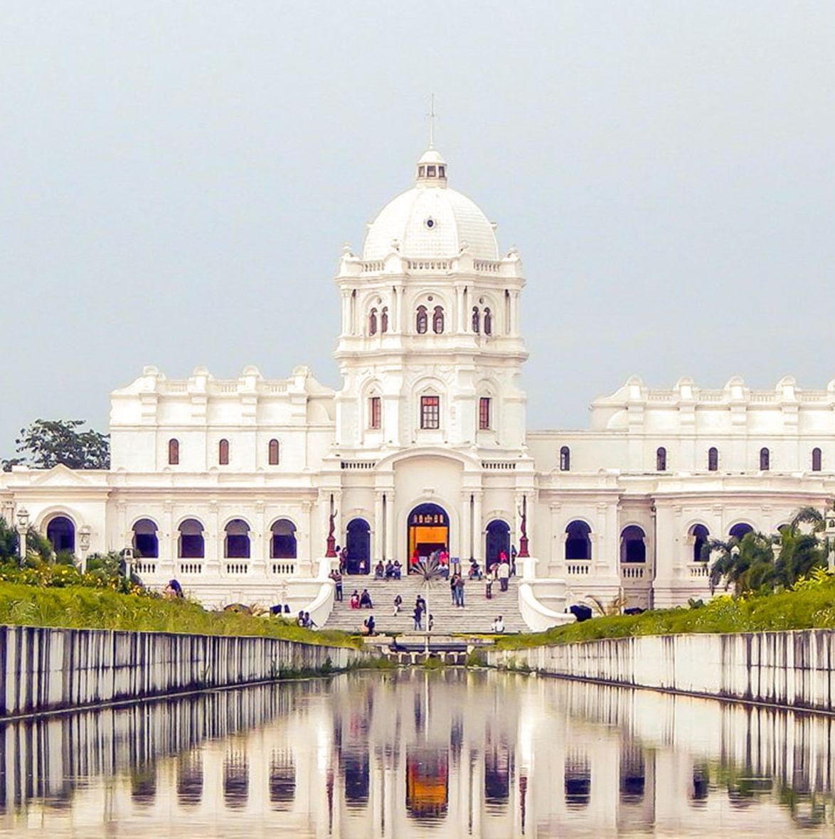 A view of the Ujjayanta Palace's facade from the pond surrounded by greenery on both sides.