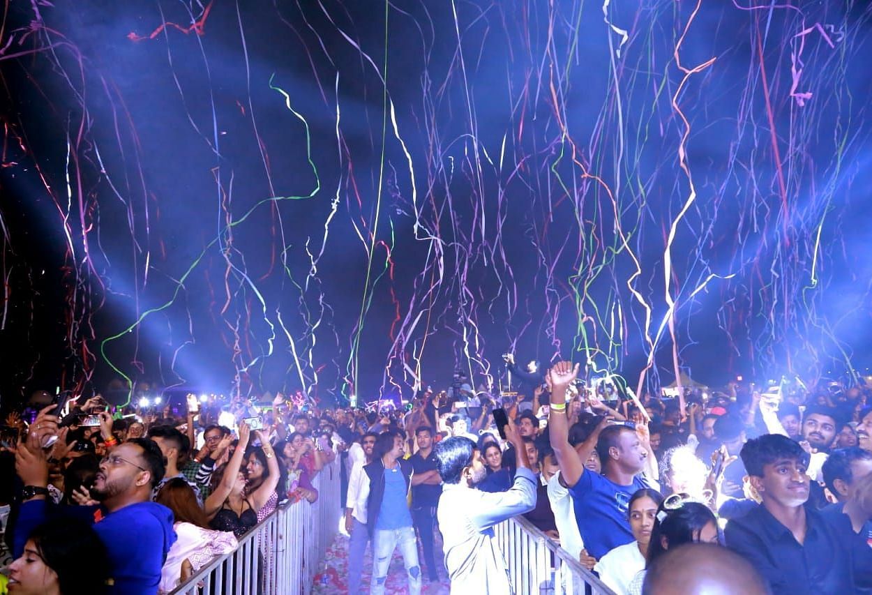 A large crowd of people is gathered in a brightly lit outdoor space at night. Many individuals have their hands raised in the air. Streams of confetti or streamers are falling from above, illuminated by stage lighting. A barrier separates the crowd from th