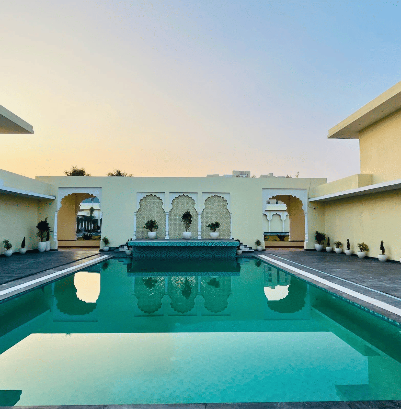 Outdoor swimming pool surrounded by arched buildings with seating areas at Beelwa Palace, Jaipur