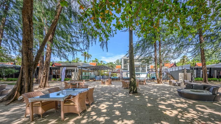 An outdoor dining area with neatly arranged tables and chairs on the sand, surrounded by tall trees - Maikhao Dream Villa Resort & Spa, Phuket