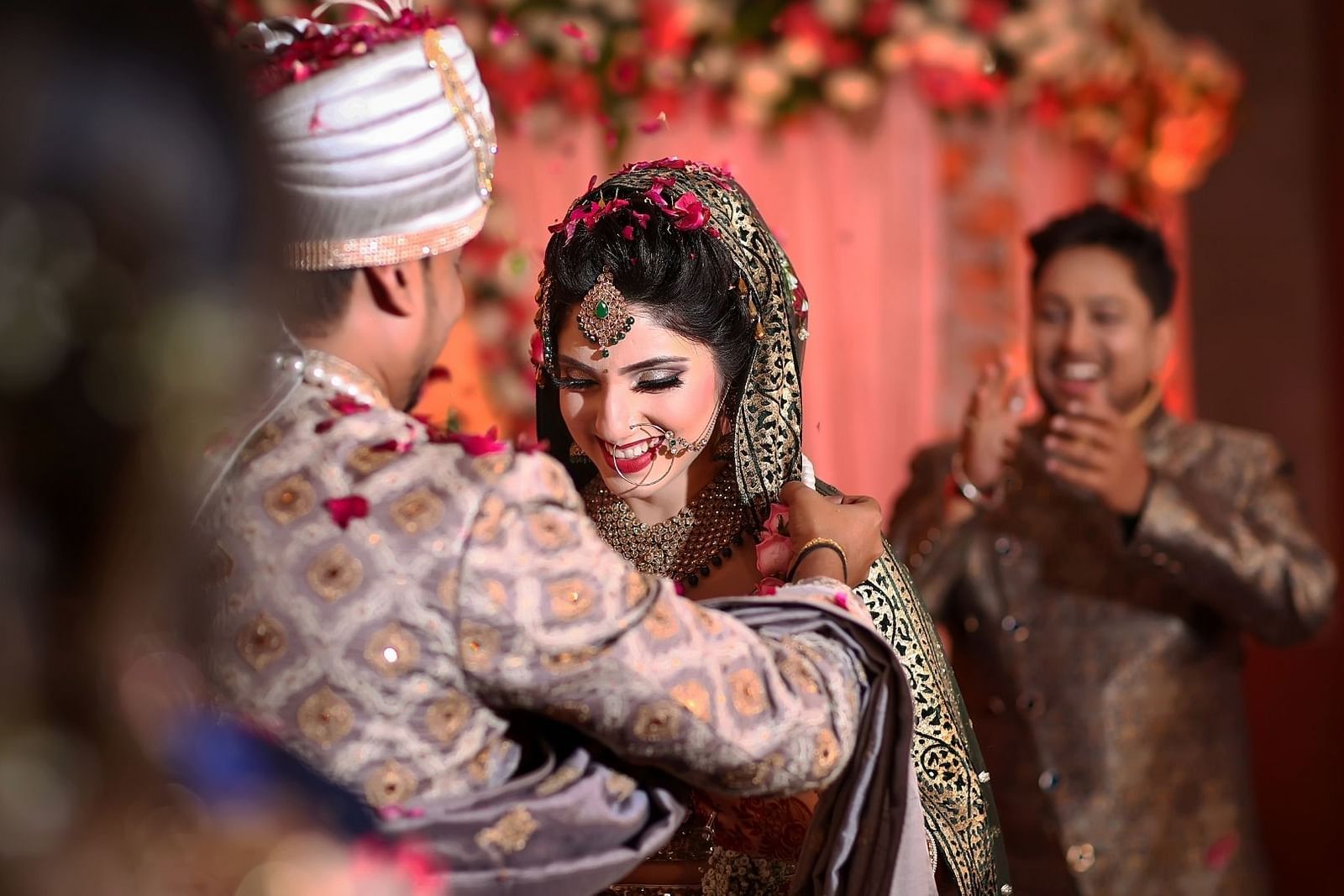 bride and groom standing together with a person clapping behind them - Rhythm Gurugram