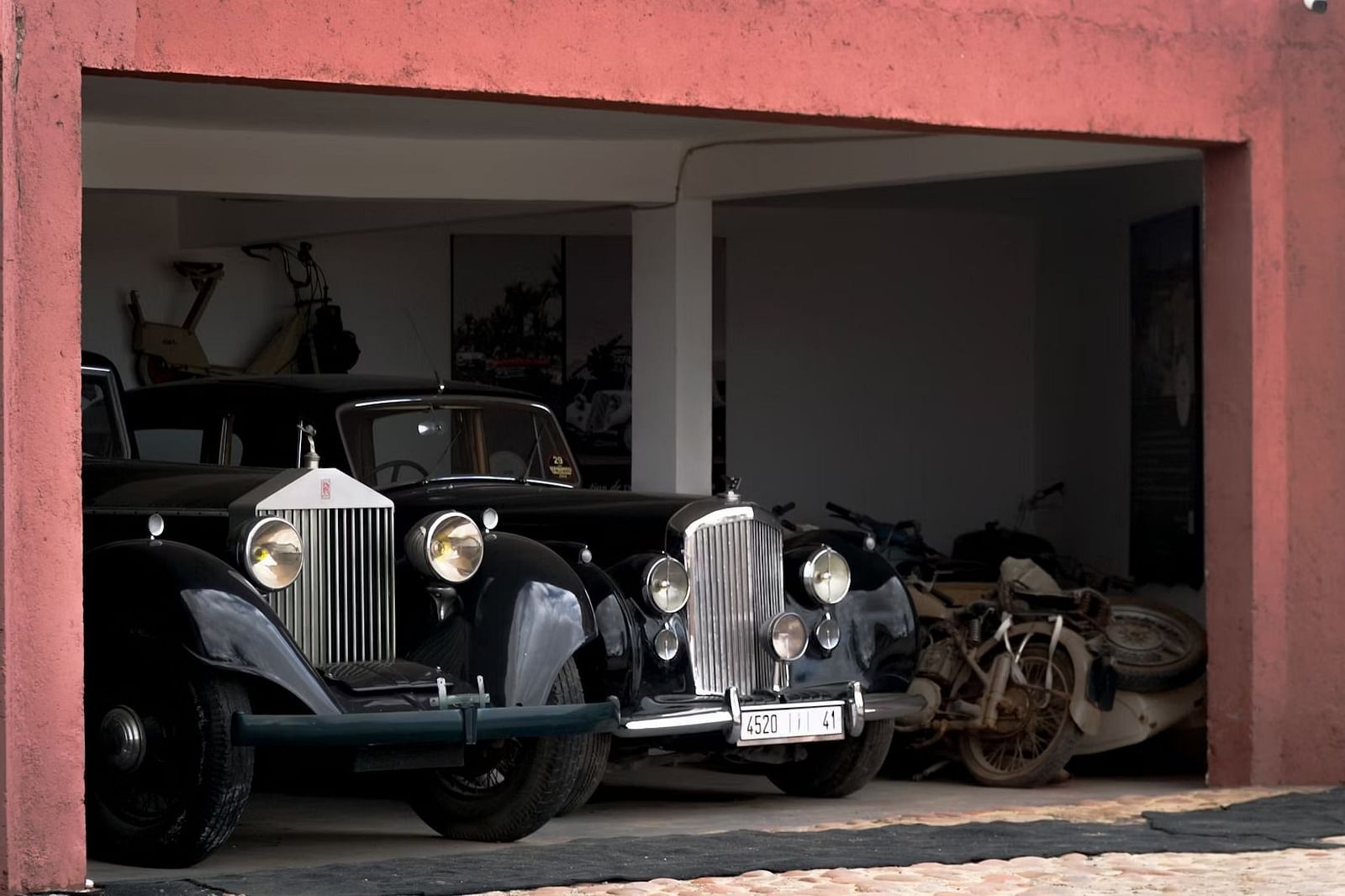 Two vintage Rolls-Royce cars parked inside a garage with old motorcycles in the background.
