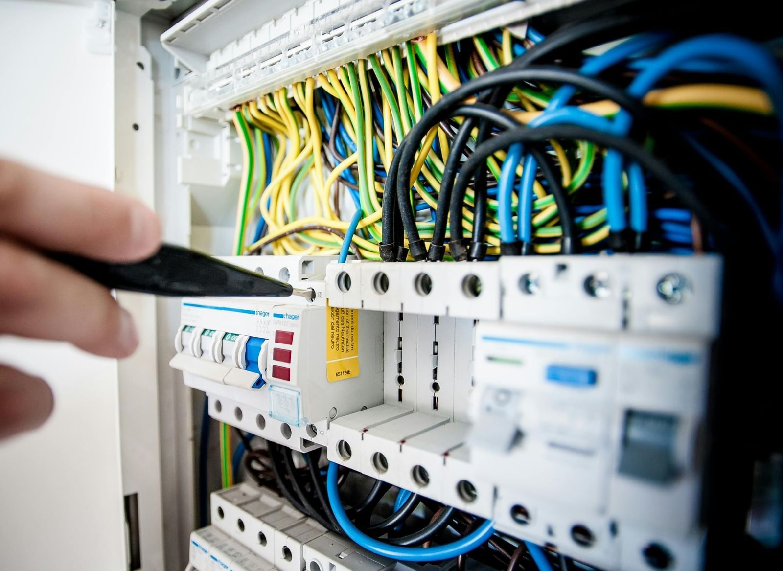 A close-up shot of an electrician working on a fuse box. The person is holding a tool in their right hand and touching a component in the electrical panel. 