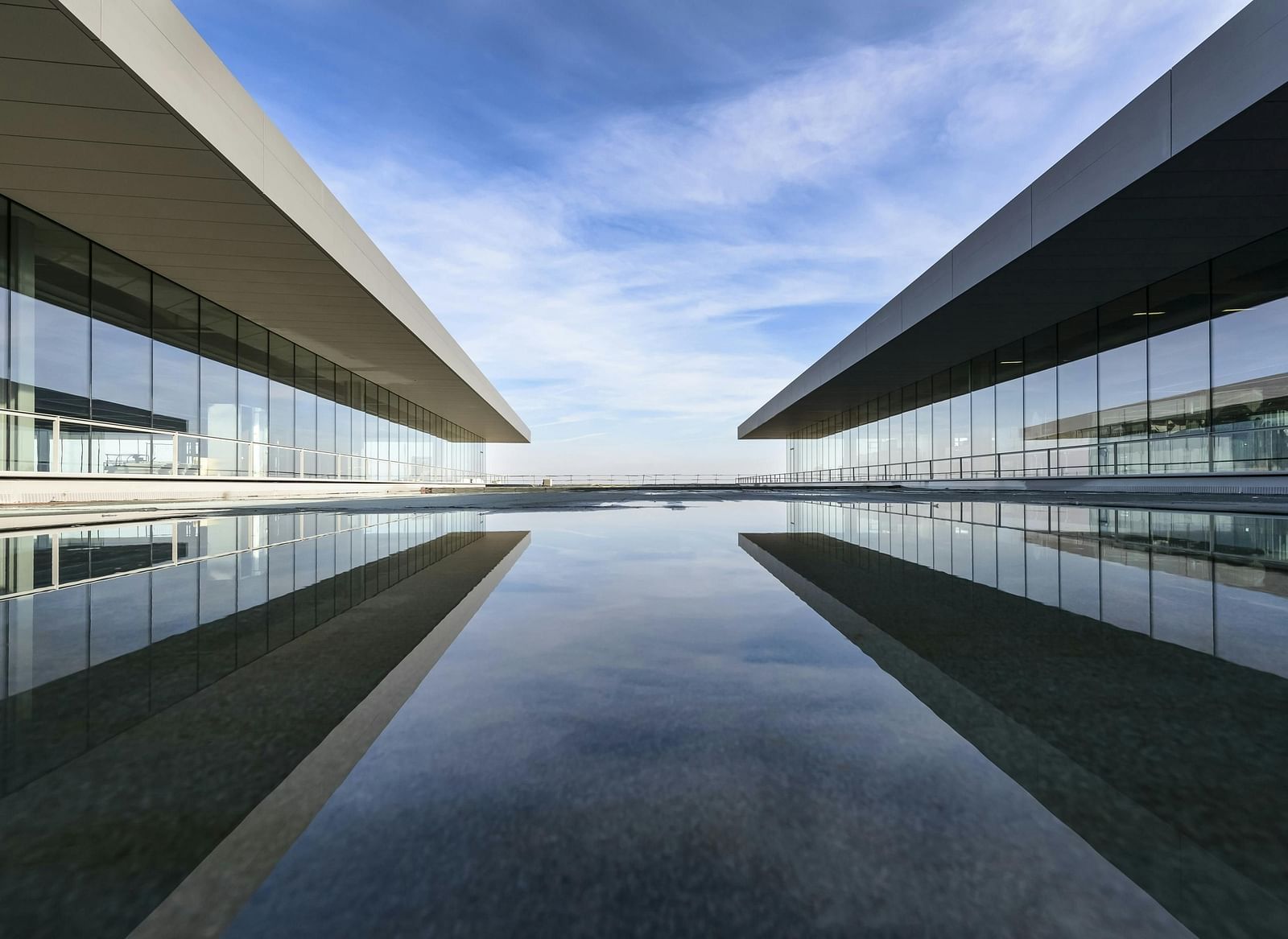 Two modern buildings with angled roofs face each other across a still body of water, creating a symmetrical reflection.
