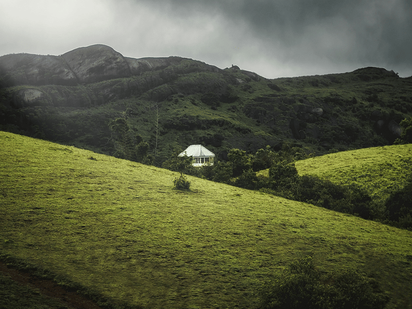 A mountain slope under a cloudy sky, indicating monsoon Vagamon weather, with a building tucked away among the trees