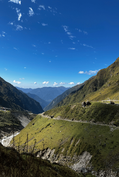 A narrow, winding road hugs the side of a steep, grassy mountain slope overlooking a deep valley under a bright blue sky.