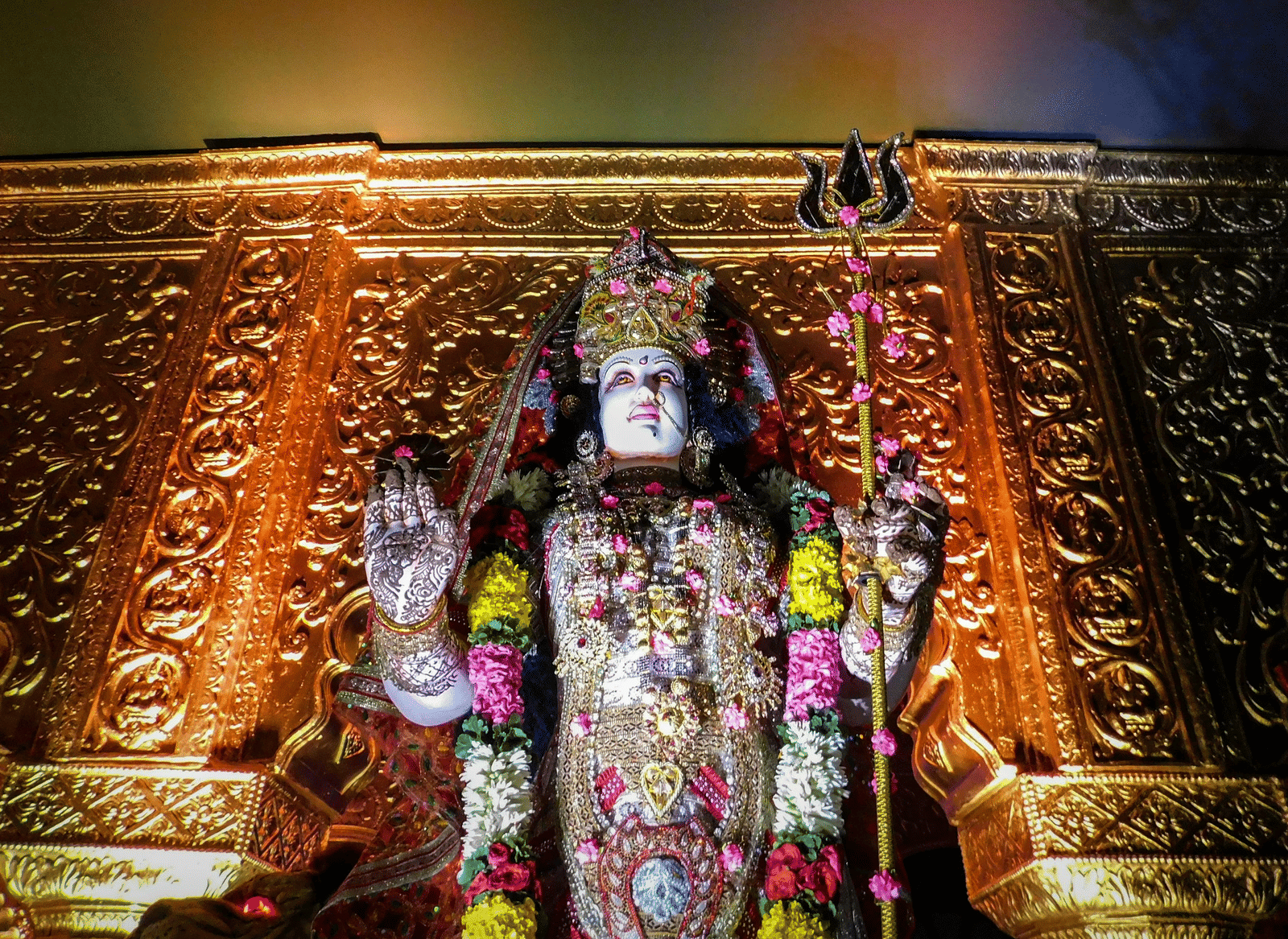 Decorated statue of  Goddess Durga with a trident and a flower garland, set against a shining golden altar.