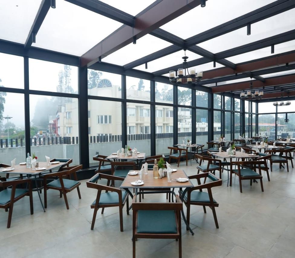 A rooftop dining area with glass walls and ceiling, offering stunning views and arranged tables and chairs for guests at The Hamuse Hotel, Kodaikanal.