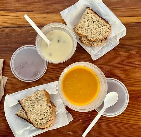 Two containers of soup, one light and one dark, sit on a wooden table next to two pieces of bread and napkins.