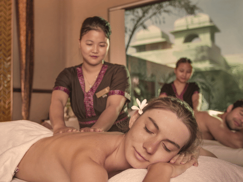 A couple receiving simultaneous back massages, with a large window showing an exotic building outdoors at Heritage Village Resorts & Spa, Manesar, one of the sought-after wellness resorts near Delhi