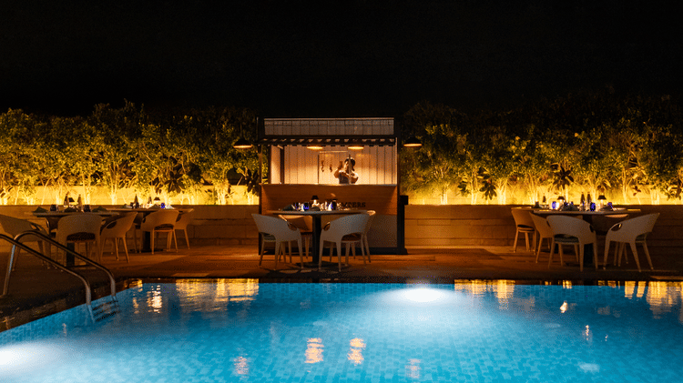 A tranquil nighttime view of the hotel's outdoor swimming pool, lit up with glowing lights, with a seating area surrounded by plants at The Residency Towers Rameswaram.