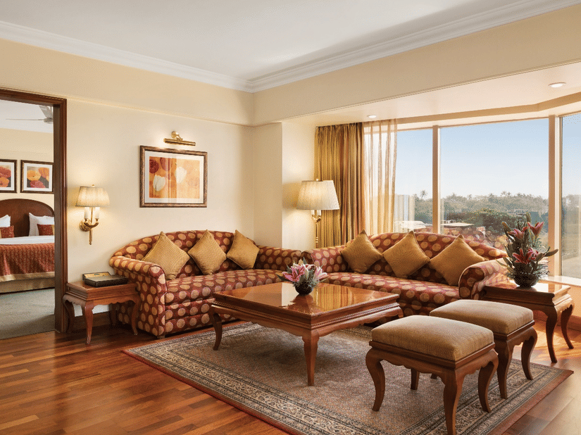 Living area of a suite with multiple sofas, a wooden centre table, lamps, and a big window with a view of the natural scenery outside.