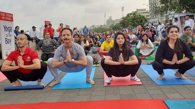 A large group of people participating in a 'Yoga by the Bay' event, practising the Malasana (garland pose) outdoors on yoga mats by the seaside promenade, with hands in prayer position.