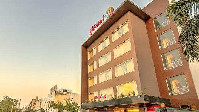 An exterior view of Hotel 91 HUDA Premier, Gurugram building with signage, entrance, parked cars, and surrounding road.