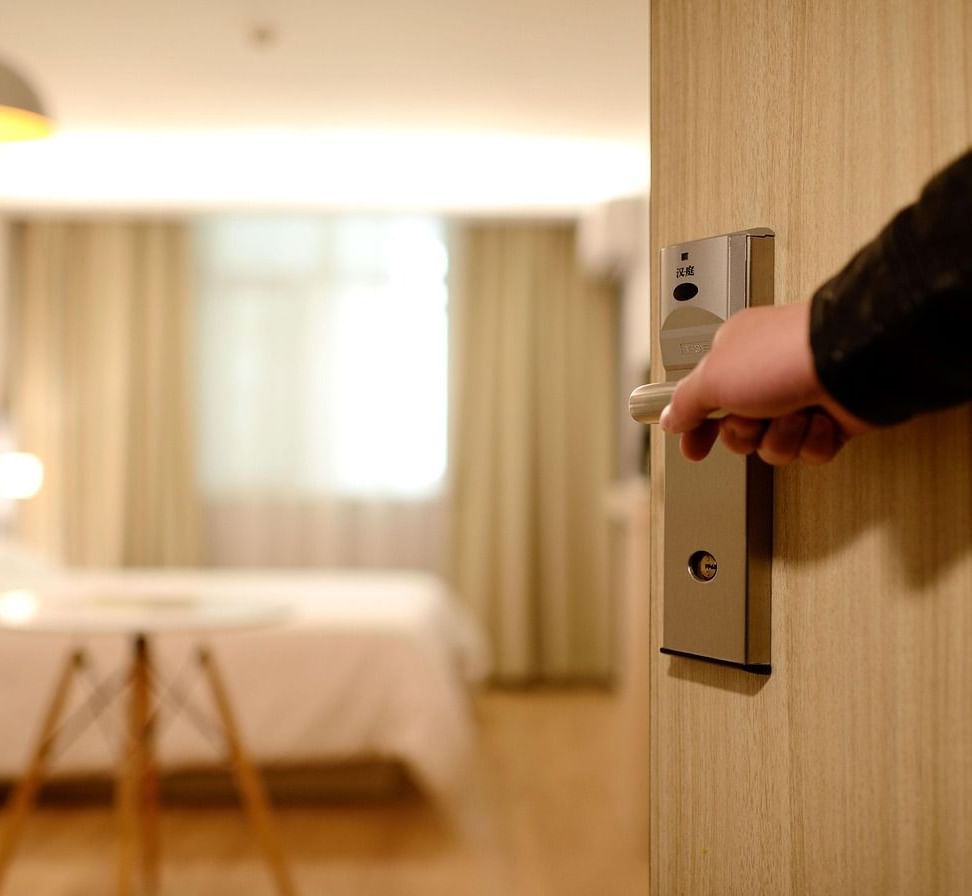 A person opening the door of a room featuring brightly-lit interiors and a double bed inside it.