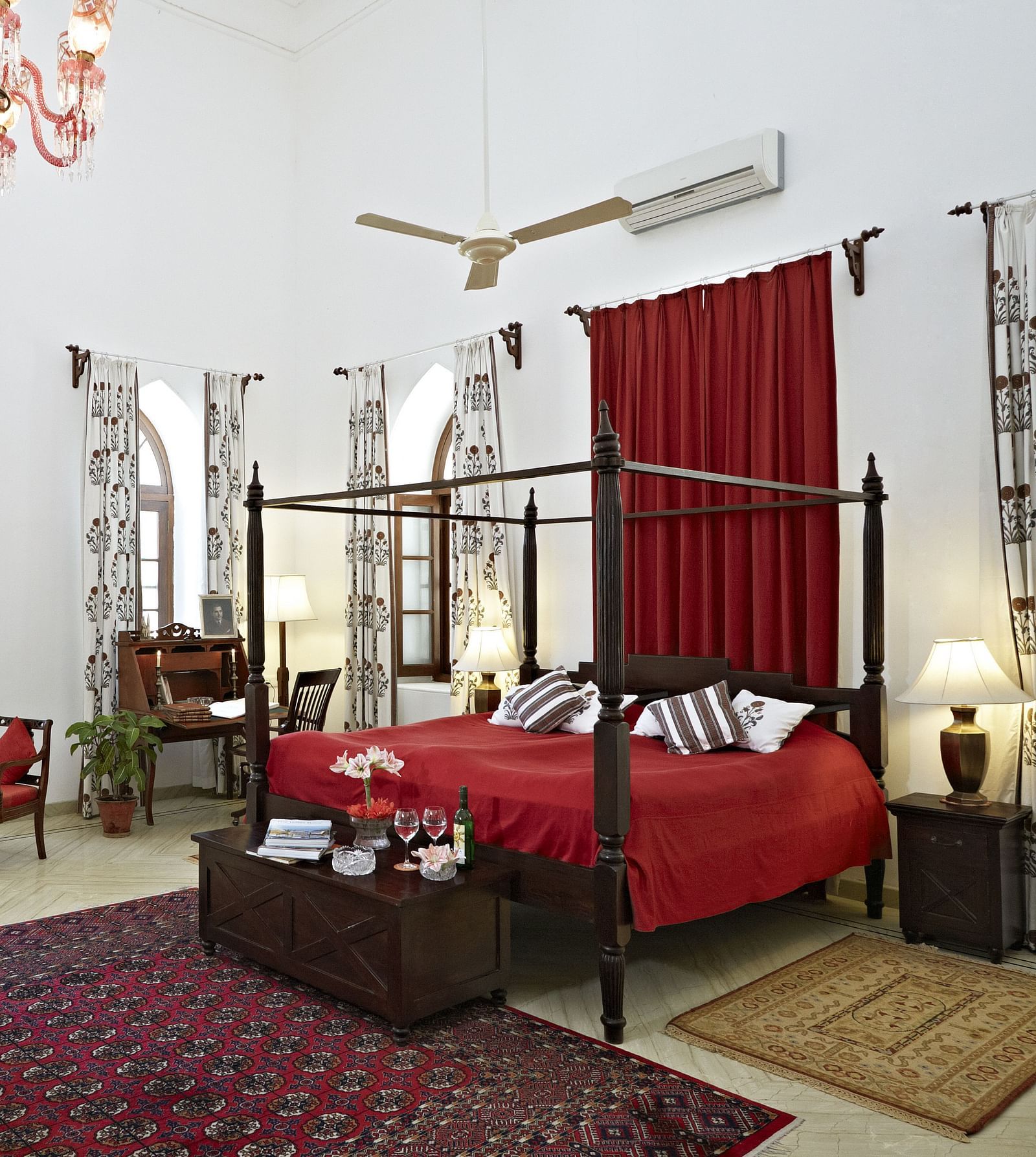 A spacious room featuring a four-poster bed with linens, seating arrangements, decorative rugs, and arched windows with draped curtains- Shahpura Bagh.