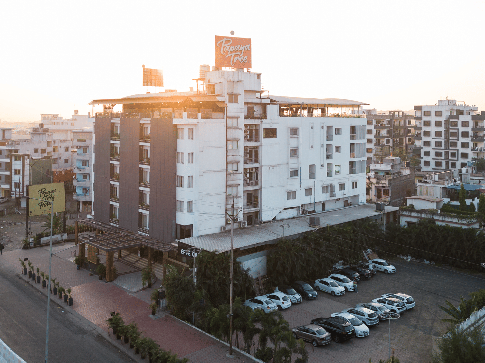A wide exterior view of Papaya Tree Hotels captured during sunset, highlighting the building’s white facade, rooftop area, and surrounding city streets with parked vehicles.