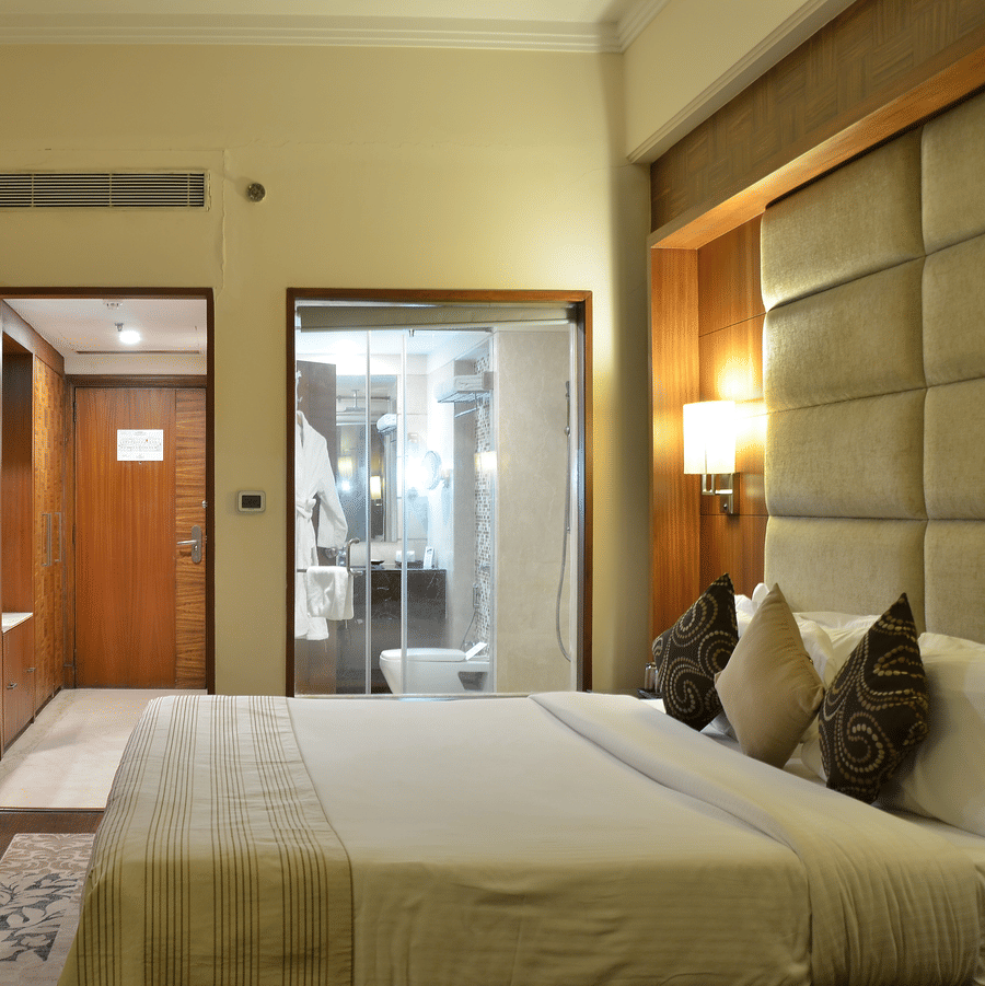 Premium Room at The Bristol Hotel, Gurgaon with a king-size bed, wall-mounted TV, a wooden wardrobe area, and a glass-enclosed bathroom with a walk-in shower.