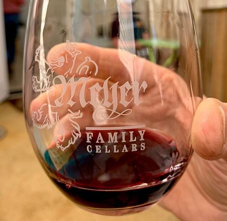 A hand holds a wine glass filled with red wine, featuring the Meyer Family Cellars logo.