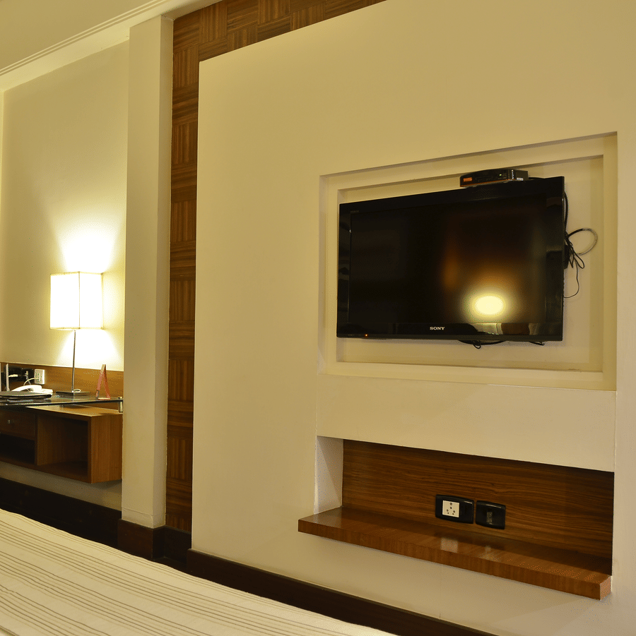 Premium Room at The Bristol Hotel, Gurgaon with a work desk and black chair, a desk lamp, a wall-mounted flat-screen TV in a wood-trimmed niche, and curtains.