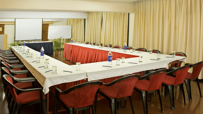 U-shaped seating inside the Mantara Conference Hall with red chairs, a projector screen, a white board, and curtains covering the windows at VITS Kamats Resort, Silvassa.