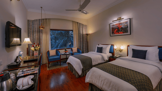A Premiere Room at The Clarks Varanasi featuring twin beds, a ceiling fan, a wall-mounted television, and a seating area at The Clarks Varanasi.