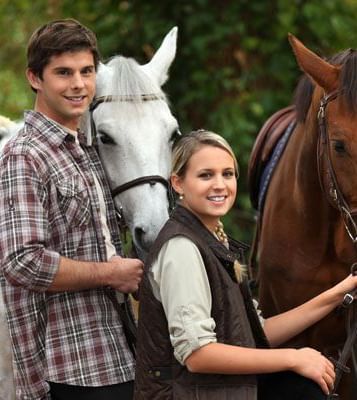 A man and a woman posing with a white and brown horse in an outdoor setting.