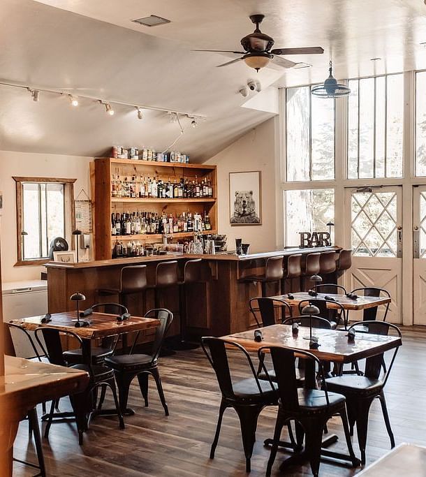 A cafe at Shaver Lake Village Hotel with wooden tables, metal chairs, large windows, and warm lighting, creating an inviting ambiance.