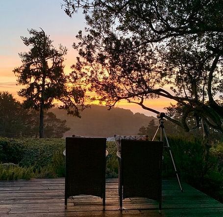 An image of a deck where two chairs are facing towards the sun set and the entire area is surrounded by trees