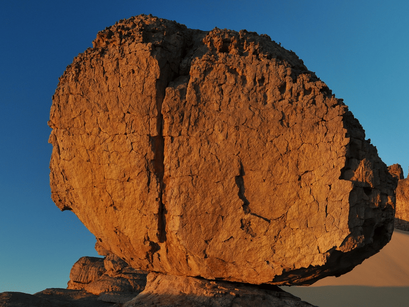 A large, rounded rock formation balancing on a narrow base in a desert landscape with sand dunes under a clear sky.