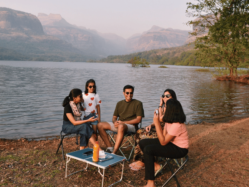 Four friends sitting in chairs by a lake enjoying a picnic, with hills and mountains in the hazy background.