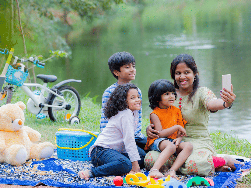 Mother taking a selfie with her three children while picnicking by a pond; a teddy bear and bike are nearby.