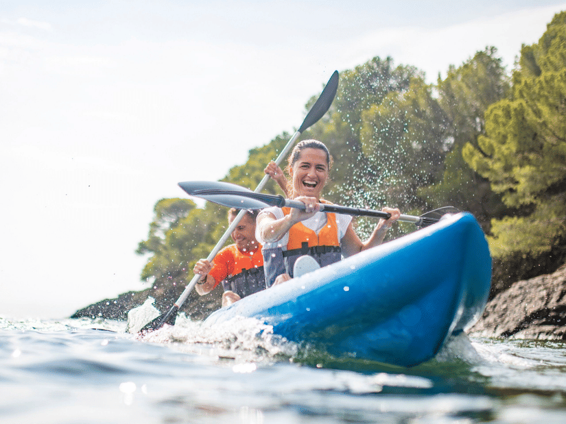 2 people smiling and paddling an inflatable blue kayak on a lake on a sunny day.